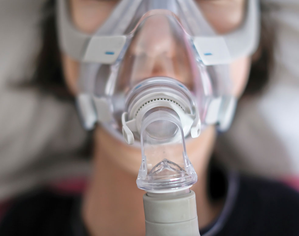 Patient wearing a CPAP breathing mask connected to medical respiratory equipment.